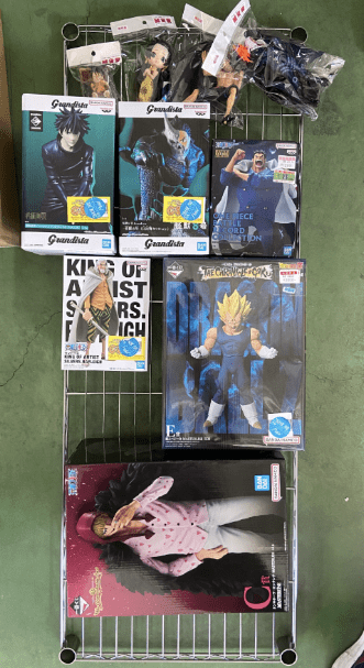 Ver Fotos Recientes 352 - Articulos Japan (post Box Japan) A metal grid shelving unit with multiple boxed action figures, anime and superhero character boxes arranged on shelves, some figures still in packaging on top shelf. Articulos Japan (Post Box Japan)