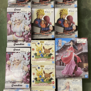 Grid of boxed anime figures on a floor shelf: Naruto-like duo holding a baby, plus other figures (white-haired heroine, Pikachu/Eevee sets, pink-dress character).
