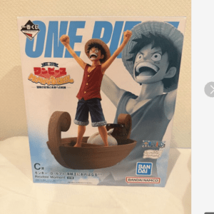 Boxed One Piece figure of Monkey D. Luffy standing on a wooden boat with arms raised, oversized blue ONE PIECE logo in the background.