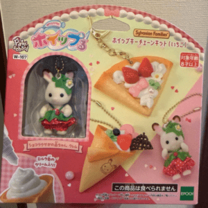 Packaging for a Sylvanian Families mini jewelry set featuring a white bunny figure with green hat and strawberry dress, plus dessert charms on pink background.