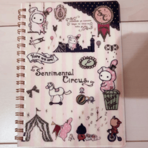 Pastel pink spiral notebook cover with Sentimental Circus characters, a carousel horse, circus tents, clock, and whimsical banners.