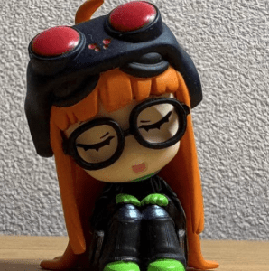 Stylized doll with orange hair, large round glasses, wearing a black helmet with red goggles, green shoes, and a black outfit, sitting on a wooden surface.
