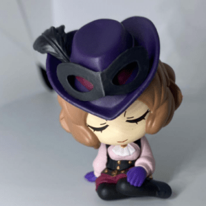 Chibi figurine wearing a purple hat with a feather and black mask, seated cross-legged with eyes closed in a meditative pose.