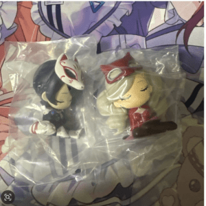 Two small chibi figurines in plastic bags on a colorful anime backdrop; left figure has dark hair and a white fox mask, right figure has light hair in a red outfit, seated with a brown prop.