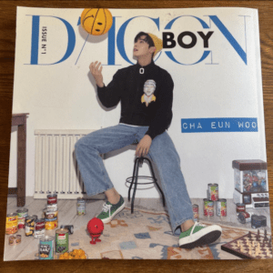 Vogue magazine cover featuring Cha Eun-woo sitting on a stool in jeans and green sneakers, surrounded by toys and canned goods. The large blue 'VOGUE' logo and 'BOY' title are visible.