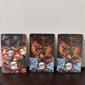 Three vertical Demon Slayer posters arranged in a row on a dark wooden surface, each featuring bold artwork of the anime with fiery and watery scenes and the series logo at the bottom left/right.