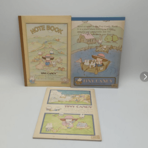 Three vintage children's books with pastel illustrations—a yellow Note Book and a blue Tiny Candy book standing upright, plus a folded pamphlet laid flat.