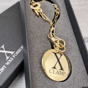Gold-tone oval pendant engraved with an X and the word 'CLAMP' on a matching chain, resting in a foam-lined jewelry box casing