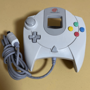 White Dreamcast game controller with four colored action buttons (Y green, B blue, X yellow, A red) and a coiled gray controller cable on a yellow surface.