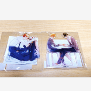 Two anime-style standee cutouts in plastic sleeves on a wooden table: left shows two characters embracing, right shows two characters sitting at a table holding hands across a panel.