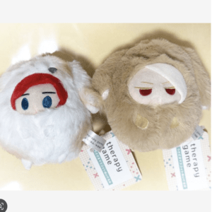Two soft stuffed plush toys, one white with red hair and blue eyes and the other tan with sleepy red eyes, lying side by side on a pale surface with 'therapy game' tags