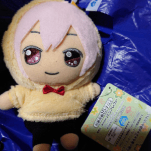 Plush toy with pink hair, yellow hoodie, red bow, black pants, lying on a blue bag with a Japanese tag attached.