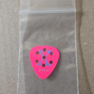Pink guitar pick with a blue geometric design and 'Japan' text, placed in a small clear bag on a brown surface.