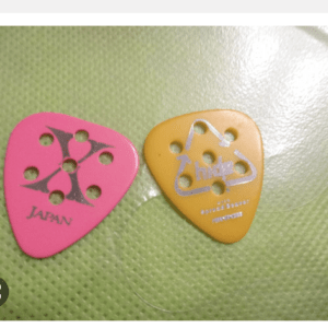 Two colored guitar picks on a green textured surface: a pink pick with a black cross pattern and 'Japan' text, and an orange pick with a white logo and several holes.