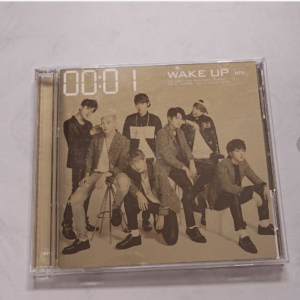 K-pop group BTS posed together for a Wake Up album cover, seven members in neutral-toned outfits in a beige background, CD case shown