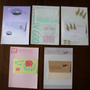 Set of six pastel-colored stationery items (envelopes and cards) with cute cartoon illustrations laid on a dark wooden surface, including purple, mint, and pink designs.