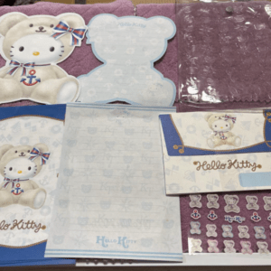 Hello Kitty-themed stationery set laid on purple fabric, including notepad, envelopes, stickers, and a bear cutout with blue ribbon.