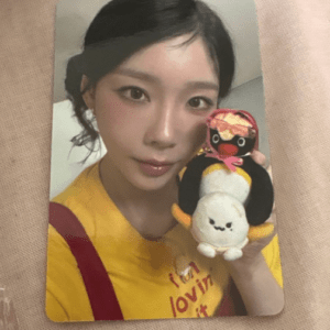 Close-up selfie of a woman in a yellow shirt holding two stuffed toys: a black-and-white penguin with a pink bow and a small white plush.