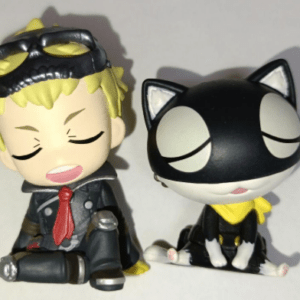 Two stylized vinyl figurines sit side by side on a plain background: a blond boy in a suit with goggles and a red tie, eyes closed as if asleep, and a black-and-white cat with a yellow scarf, also eyes closed.