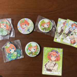 Assorted anime character merch on a wooden table: round pins, keychains, and packaged items featuring a red-haired girl with a green bow.