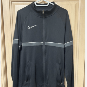 Black Nike zip-up athletic jacket on a hanger, with a light gray reflective band across the chest and sleeves and front pockets.