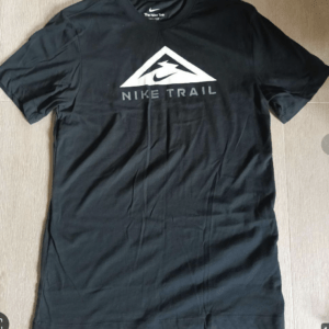 Black Nike Trail T-shirt with white triangular logo and 'NIKE TRAIL' text centered on chest, short sleeves, laid flat on a wooden floor.