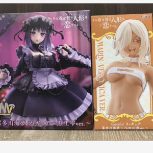 Two boxed anime figures on a wooden surface: a purple gothic-dress character on the left and an orange box with a white-haired figure on the right.