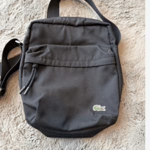 Small black backpack with front zip pocket on a beige fuzzy surface, featuring a Lacoste crocodile logo.  Articulos Japan (Post Box Japan)