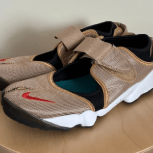 Pair of tan mesh sneakers with Velcro straps and a red Nike swoosh on a white sole, placed on a wooden table.