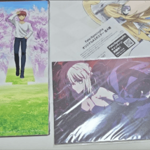 Three anime posters laid on a table: left Fate/stay night with a boy under pink blossom trees; top-right partial character art; bottom-center close-up of a pale-haired character.