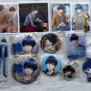 Collection of blue‑haired anime character merch: pins, badges, keychains, acrylic standee, and cards arranged on a display.
