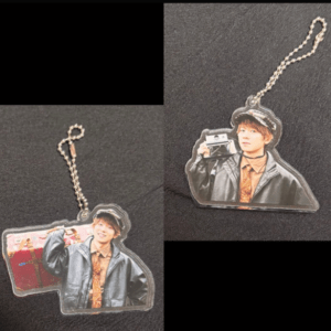 Two acrylic keychains show a man in a leather jacket and hat holding a vintage camera, photographed from different angles on a dark surface.