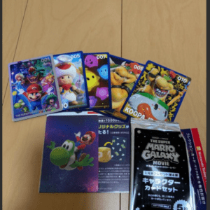 Fanned-out Super Mario trading cards on a wooden floor, featuring Mario, Yoshi, and Bowser, with a Mario Galaxy Movie card pack nearby.