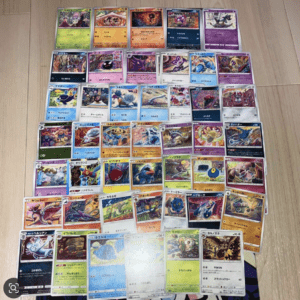 Grid of colorful Pokémon trading cards spread out on a wooden floor, arranged in rows and columns like a collection display.
