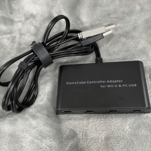 GameCube Controller Adapter for Wii U and PC USB with attached USB cable on a gray fabric surface.