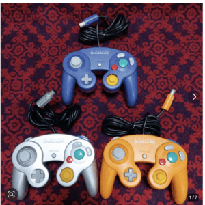 Three Nintendo GameCube controllers (blue, silver, and orange) with wired cables on a red patterned surface.