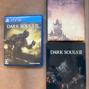 PS4 Dark Souls III game case with armored knight on the cover, placed beside two Dark Souls III art books on a wooden surface.