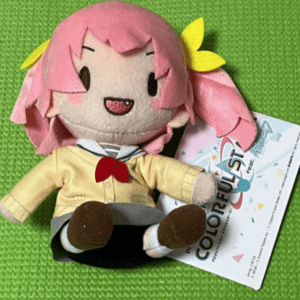 Plush toy with pink hair, yellow bow, and yellow jacket sits on a bright green, textured surface with a tag attached.