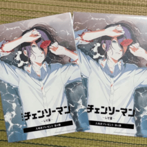 Two paperback manga volumes of Chainsaw Man laid side by side on a woven surface, showing a man in a light shirt shielding his eyes with one hand and a red flower near his head/shoulder. The title is in Japanese on the cover.