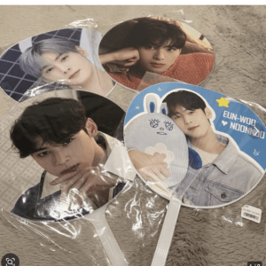 Round handheld fans showing close-up photos of a male K-pop idol, arranged on a beige rug with plastic wrapping still on the items.99