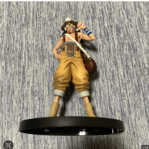 Poseable pirate action figure in yellow pants, brown belt with metal buckle, hat, goggles, and blue armband, standing on a round black display base against a striped backdrop.