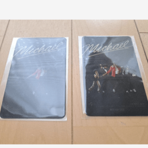 Two Michael Jackson posters in plastic sleeves on a wooden floor; left has a blue cover, right shows MJ performing.