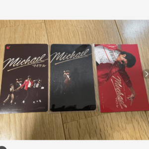 Three Michael Jackson memorabilia cards laid on a wooden floor: brown, black, and red designs featuring MJ various poses and group image.