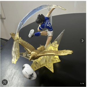 Decorative trophy showing a blue-clad soccer player leaping to kick a ball through a translucent arced ring, with a gold eagle and star base.  Articulos Japan (Post Box Japan)