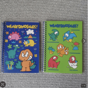 Two spiral-bound dinosaur-themed notebooks on a gray carpet: a blue cover on the left and a lime-green cover on the right, both with cartoon dinosaurs and the title 'We Are Dinosaurs!'