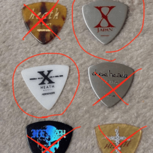 Six guitar picks laid out on a carpet; four picks are crossed out with red marks, two remain unmarked (top-right silver X JAPAN and middle-left white X HEATH).