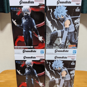 Four Grandista anime figure boxes arranged in a 2x2 stack on a wooden table, featuring two different characters on the box art with a silver-haired figure and a blue-haired figure.