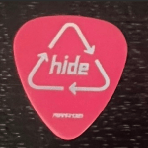 Pink guitar pick with a white recycling-triangle design and the word 'Hide' centered on it; small, illegible branding text at the bottom.