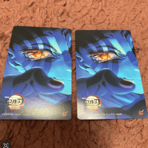 Two identical blue anime trading cards laid on a brown carpet, each showing a close-up of a face with an orange eye visually emphasized in the center of the image.