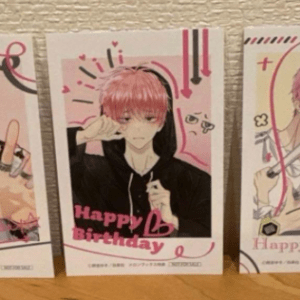 Three pink-themed birthday cards with anime-style characters on a shelf; center card features a pink-haired teen in a black hoodie with 'Happy Birthday' text.